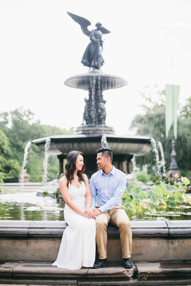 Asian couple smiling and holding hands while sitting on the edge of Bethesda fountain in Central Park, New York City USA