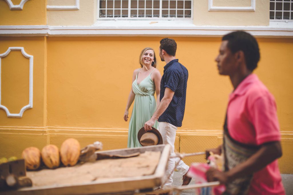 A couple walks down a street in Cartagena as a produce vendor walks by