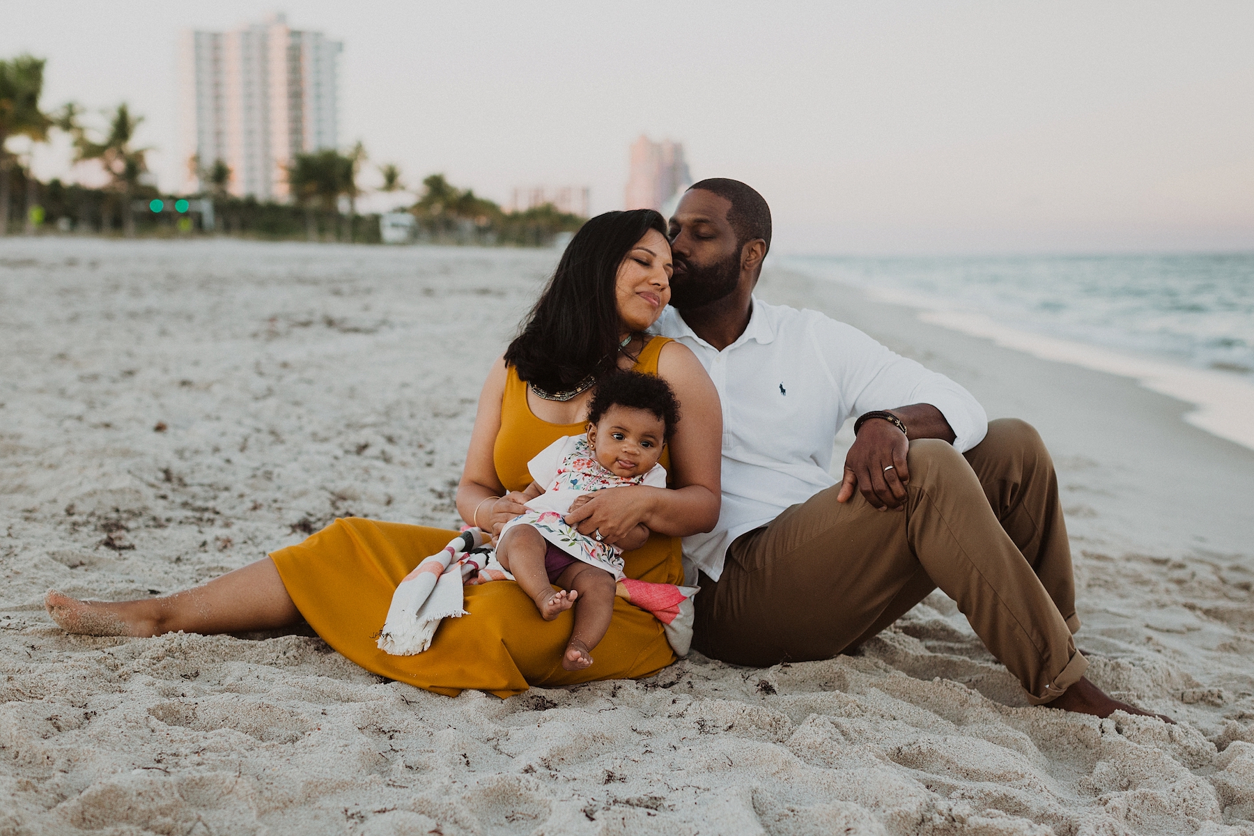 A man kisses the side of his wife's forehead while they sit on the beach with their baby in Ft Lauderdale, USA.
