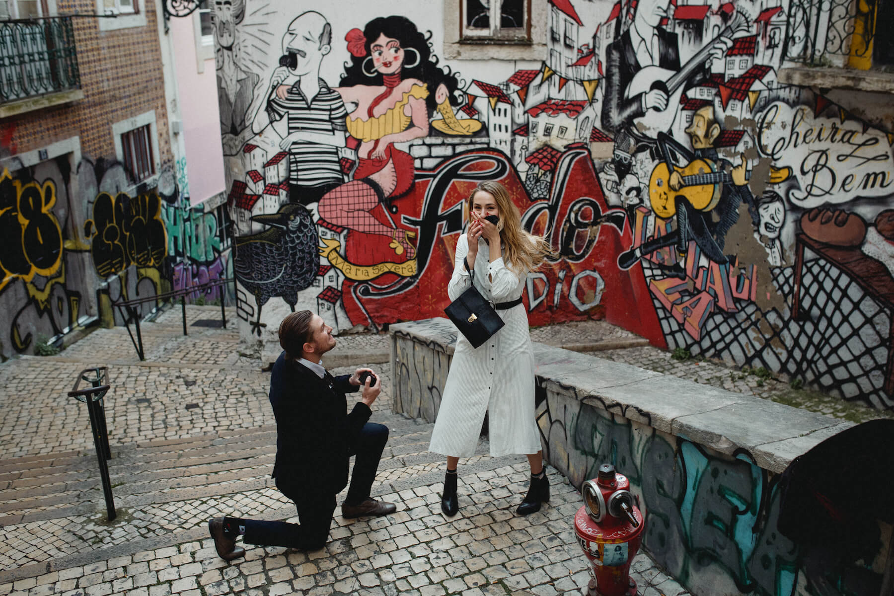 Man is on one knee proposing to the woman, who looks surprised. They are standing in front of a colourful wall in Lisbon.