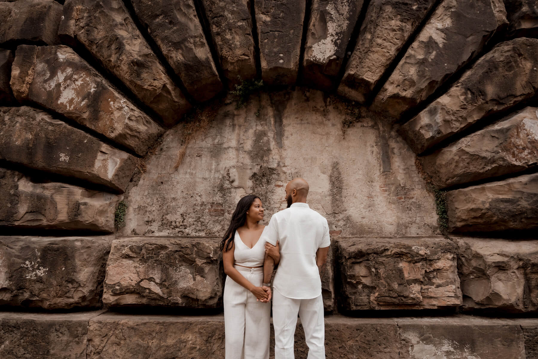 A couple sits on the ledge of a bridge laughing together in Florence, Italy while on a pre-wedding trip photoshoot for Flytographer.