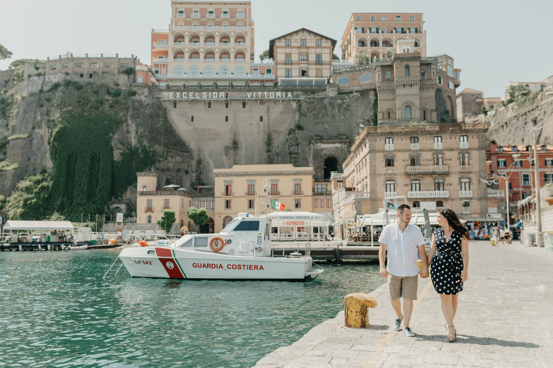 A couple walks along the sidewalk next to the water in Marina Piccola in Sorrento, Italy while on a pre-wedding photoshoot with Flytographer.